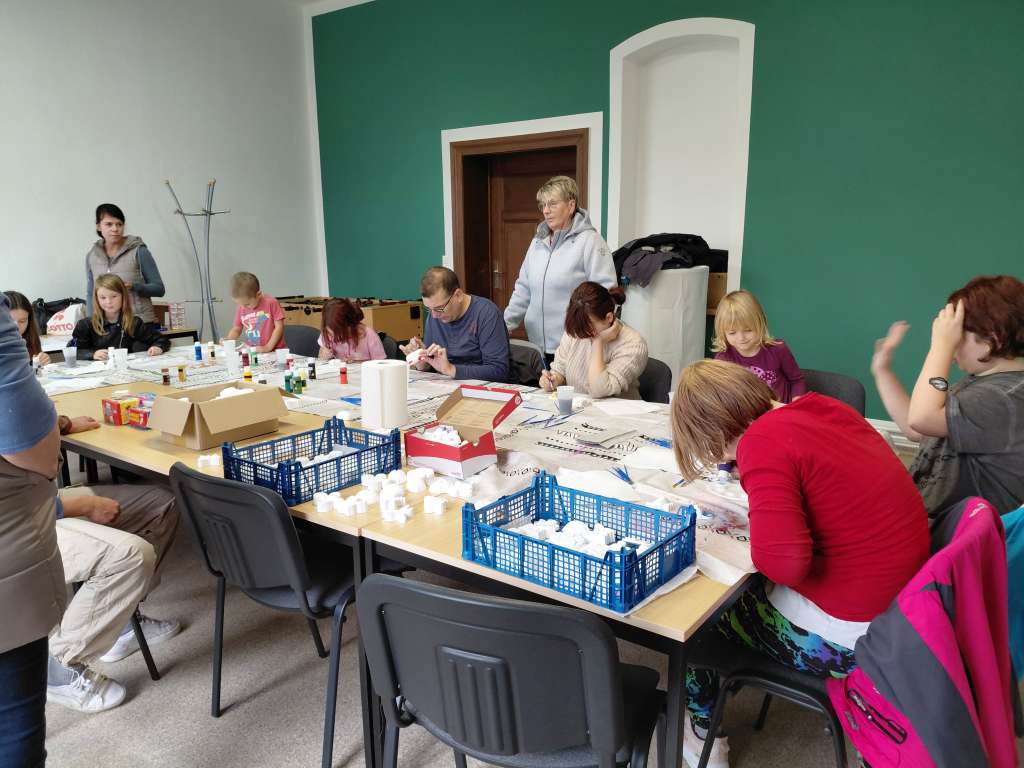 Auf dem Bild sind zwölf Menschen verschiedenster Altersgruppen zu sehen. Die meisten von ihnen arbeiten konzentriert und bemalen kleine Tonfiguren mit großer Sorgfalt.