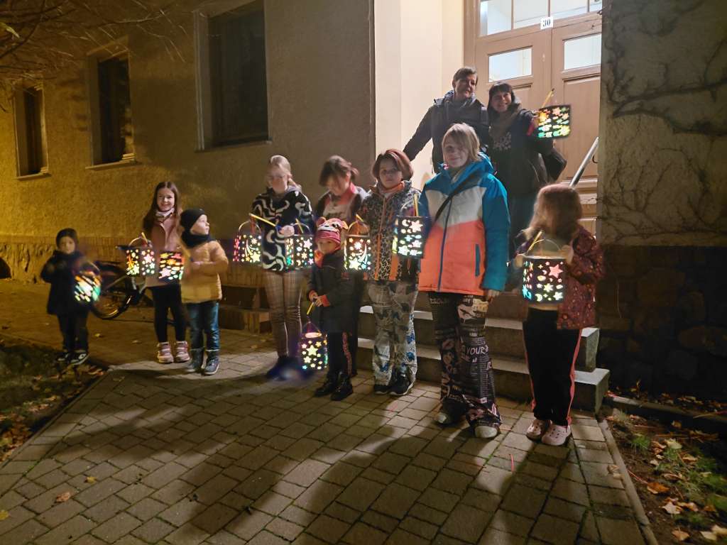 9 Kinder stehen mit ihren leuchtenden Laternen stehen fröhlich vor einer Tür. Im Türrahmen stehen 2 Erwachsene mit einer Laterne.