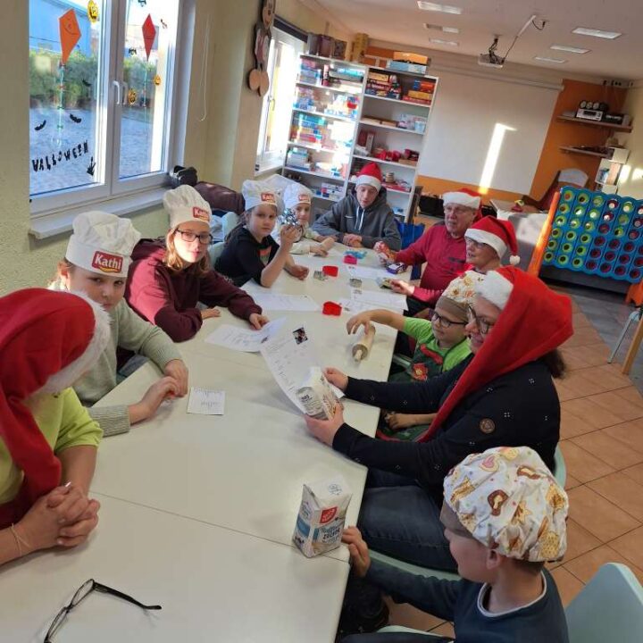 An einem langen Tisch sitzen sieben Kinder und vier Erwachsene. Alle tragen Weihnachts- oder Kochmützen und schauen sich gemeinsam verschiedene Rezepte an. Die Stimmung ist fröhlich und voller Vorfreude aufs gemeinsame Kochen und Backen.