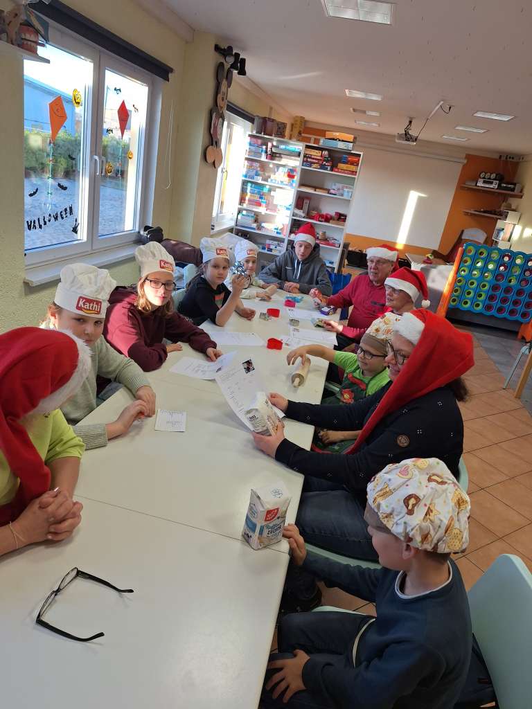 An einem langen Tisch sitzen sieben Kinder und vier Erwachsene. Alle tragen Weihnachts- oder Kochmützen und schauen sich gemeinsam verschiedene Rezepte an. Die Stimmung ist fröhlich und voller Vorfreude aufs gemeinsame Kochen und Backen.
