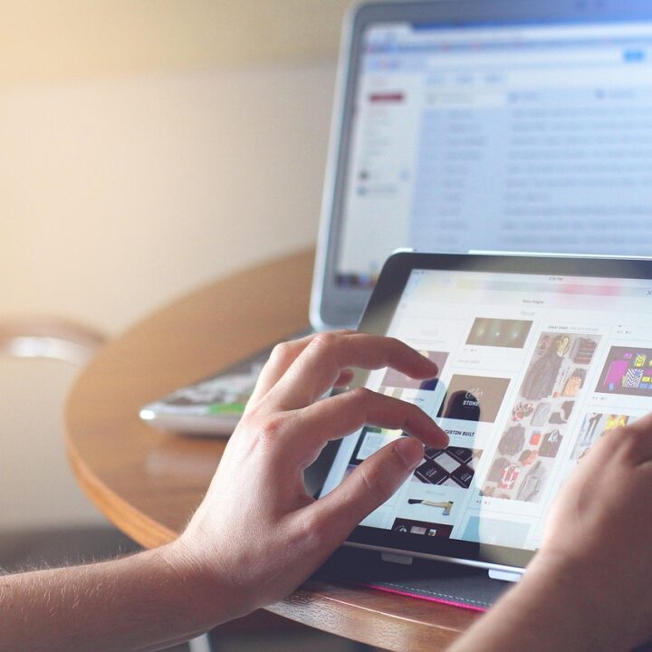 Hand scrollt über ein Tablet. Im Hintergrund steht ein Laptop